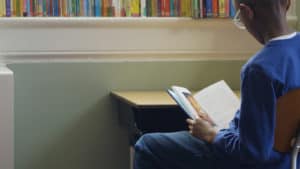 Black schoolboy in blue sweatshirt, sitting at desk, reading book